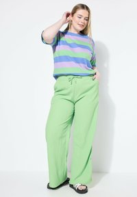 Gestreepte t-shirt in paars, groen en lavendel; losse pasvorm. Lichtgroene, gestructureerde broek met een elastische tailleband en een trekkoord. Zwarte sandalen.