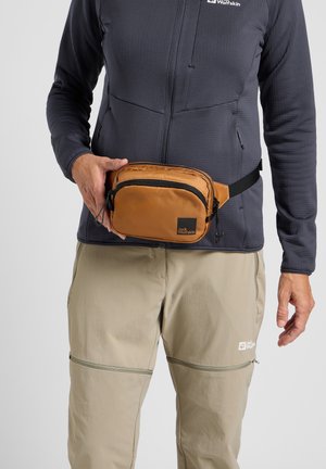 Riñonera tan hecha de un tejido duradero, con un bolsillo negro con cremallera y un logo. Combinada con una chaqueta gris y pantalones beige claros.