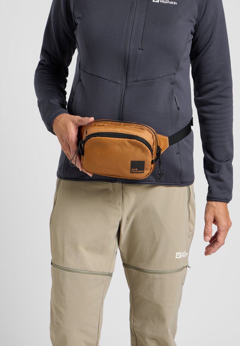 Riñonera tan hecha de un tejido duradero, con un bolsillo negro con cremallera y un logo. Combinada con una chaqueta gris y pantalones beige claros.
