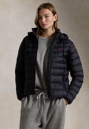 Ung kvinde iført en sort quiltet jakke over en grå sweatshirt og grå joggingbukser med snørre, stående med hænderne i lommerne.