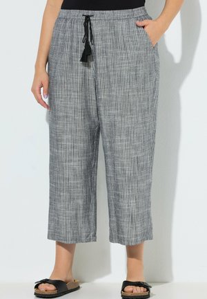 Kobieta w szaro-białych, pionowo paskowanych spodniach materiałowych z czarnym troczkiem i czarnych klapkach na płaskim obcasie z kokardką.