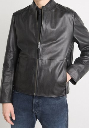 Mann trägt eine schwarze Lederjacke mit Reißverschluss über einem schwarzen Hemd und Krawatte, kombiniert mit blauen Jeans, mit einer Hand in der Tasche.