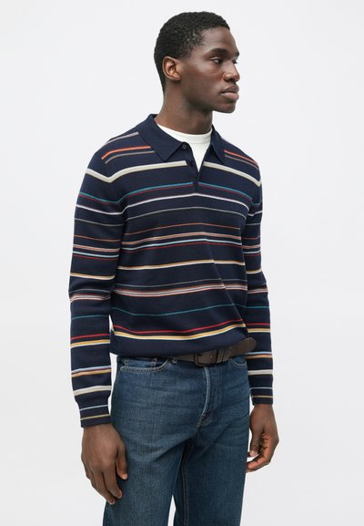 Pruhovaný námornícky modrý pulóver z pleteniny s viacfarebnými horizontálnymi pruhmi. Má golierový výstrih a gombíky. Skombinovaný s modrými džínsami.