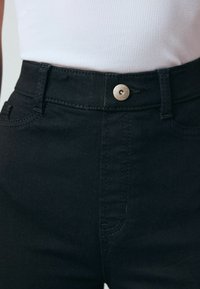 Black high-waisted jeans with a metal button closure, featuring a smooth texture and contrast stitching along the pockets and seams.