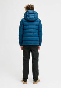 Garçon aux cheveux bouclés portant une doudoune à capuche bleue, un pantalon cargo noir et des bottes beiges, se tenant de dos sur fond blanc.