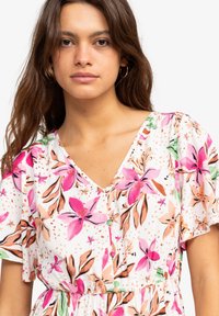 Bloemjurk met een witte achtergrond, roze en groene bloemenpatronen, een V-halslijn en korte fladdermouwen. Geweven stofdetail.