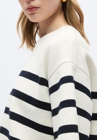 Κοντινό πλάνο ενός ατόμου που φοράει κρεμ πουλόβερ με τολμηρές μαύρες οριζόντιες ρίγες και χρυσό κρίκο σκουλαρίκι, με τα μαλλιά κάτω στον ώμο.