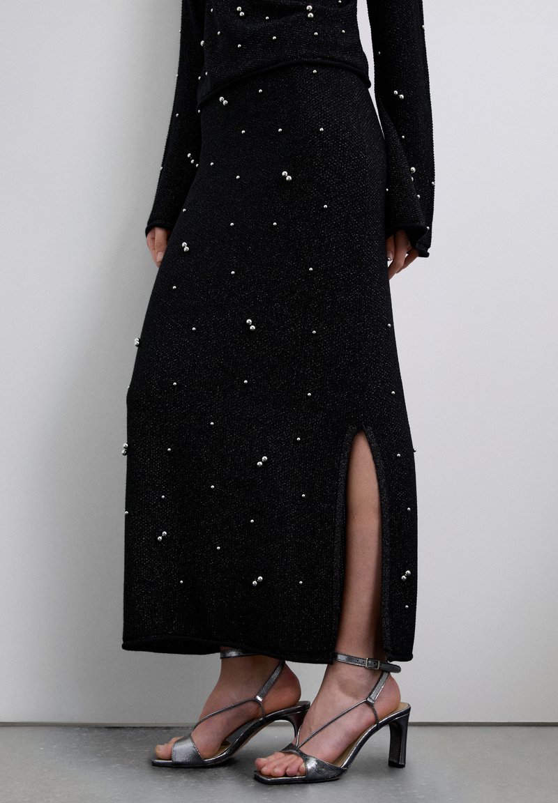 Vestido negro de manga larga con hilos metálicos y detalles de perlas dispersas. Cuenta con una abertura lateral y se combina con sandalias plateadas de tacón.