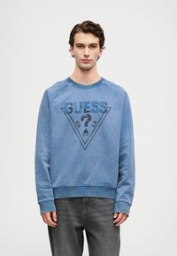 Joven vistiendo una sudadera azul de Guess con un logo triangular y un signo de interrogación, combinada con jeans gris oscuro, de pie contra un fondo sencillo.