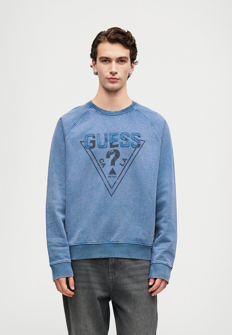 Joven vistiendo una sudadera azul de Guess con un logo triangular y un signo de interrogación, combinada con jeans gris oscuro, de pie contra un fondo sencillo.