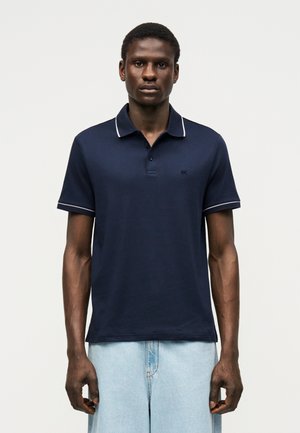 Mann steht mit Blick nach vorne, trägt ein marineblaues Poloshirt mit weißer Einfassung und hellblaue Jeans vor einem schlichten hellen Hintergrund.