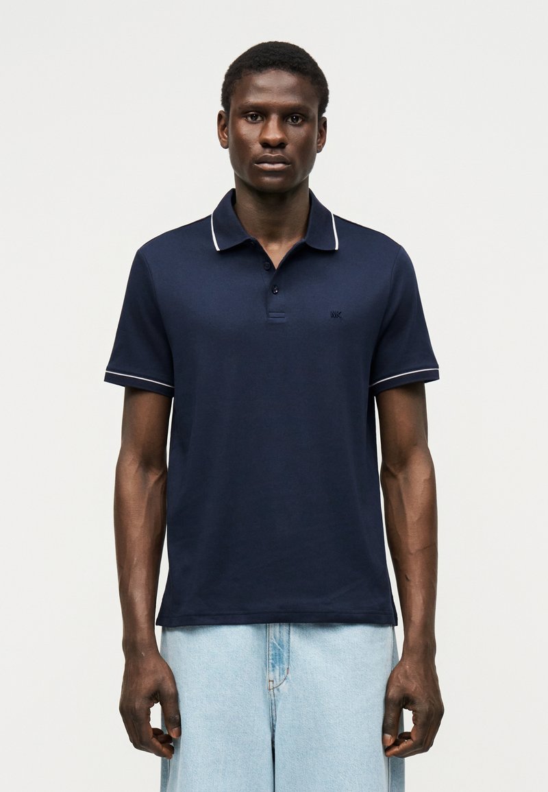 Homme debout face à l'avant portant un polo bleu marine avec une bordure blanche et un jean bleu clair sur un fond clair uni.