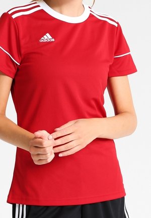 Persona che indossa una maglietta sportiva rossa Adidas con strisce bianche sulle spalle e pantaloncini neri, mani giunte davanti.