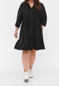 Robe noire à manches longues, à boutons, en tissu doux et texturé, avec une jupe évasée et un décolleté en V, associée à des baskets blanches.