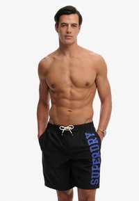 Homme musclé aux cheveux courts et foncés, portant un short de bain noir avec le texte bleu "SUPERDRY", debout les mains dans les poches sur un fond blanc.