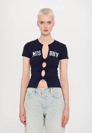 Marineblå kortærmet top med udskæringsdetaljer, tre sløjfer på forsiden og teksten "MIS BHV". Båret med lyseblå mønstrede højhvide jeans.