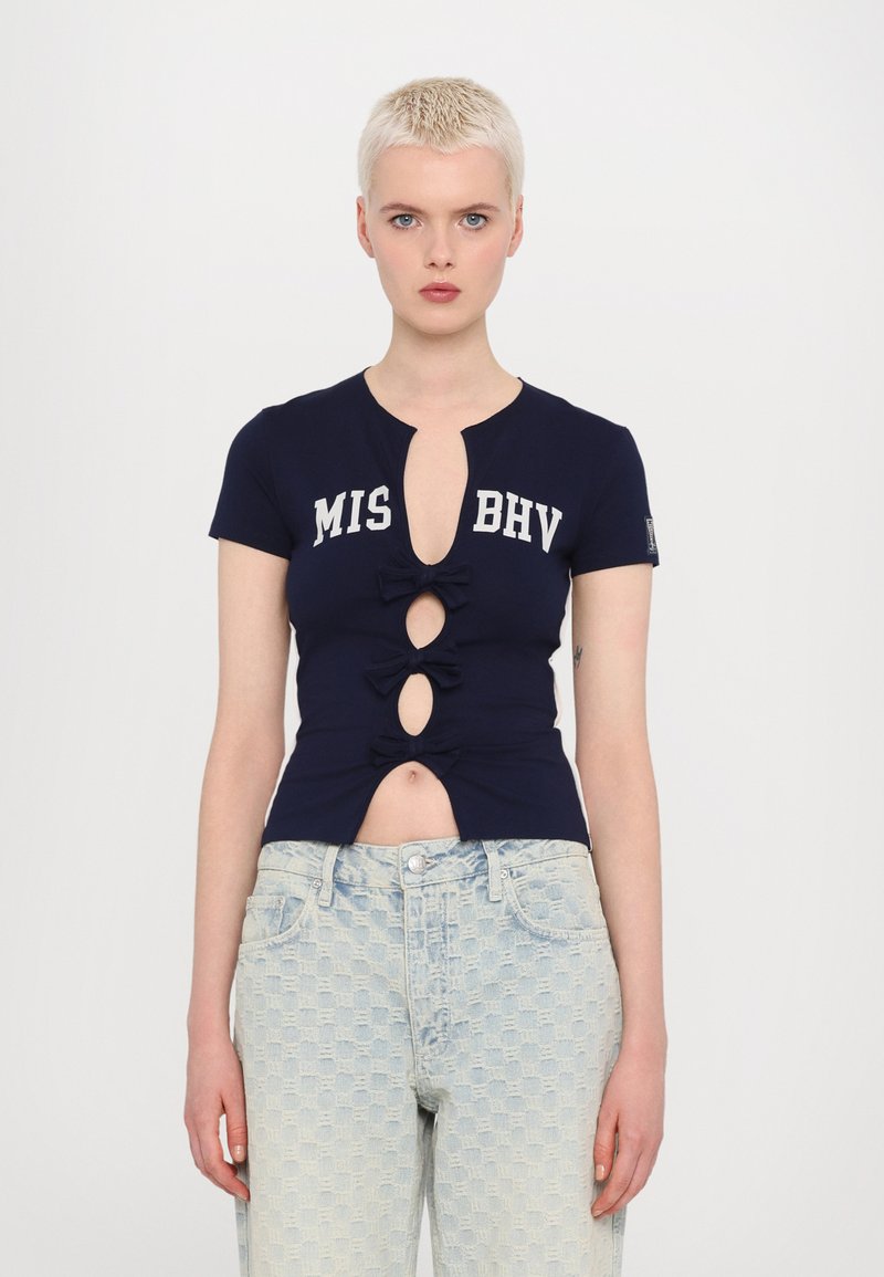 Navy short-sleeve top with cut-out detailing, three bows on the front, and "MIS BHV" text. Worn with light blue patterned high-waisted jeans.