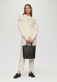 Strickpullover in hellbeige mit schulterfreiem Design, kombiniert mit cremefarbenen Hosen. Schwarze Ledertasche mit Krokodilstruktur und einer kleinen Tasche.