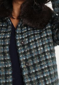 Vävd jacka i ett grönt, blått och grått houndstooth-mönster. Har en mörk pälskrage och svarta knappar. Strukturerad, varm tyg.