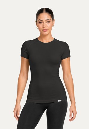 Femme aux cheveux foncés attachés, portant un t-shirt noir ajusté à manches courtes et des leggings noirs, debout les bras détendus le long du corps.