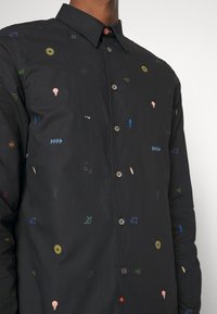 Chemise noire à boutons avec de petits symboles et formes abstraits colorés dispersés sur le tissu, portée par une personne.