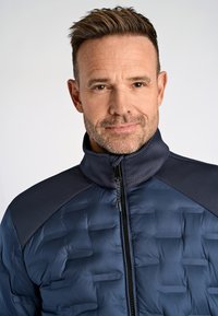 Marinblå vadderad jacka med texturerade axlar och hög krage, med en framsida som har dragkedja och ett mönster av upphöjda horisontella linjer.