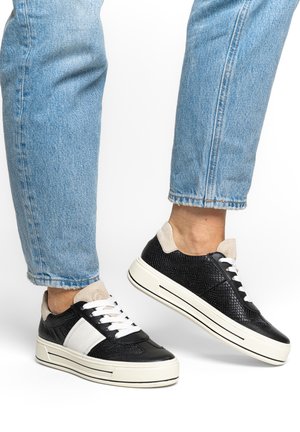 Persona con zapatillas bajas en blanco y negro con detalles texturizados y vaqueros recortados azul claro sobre un fondo blanco.