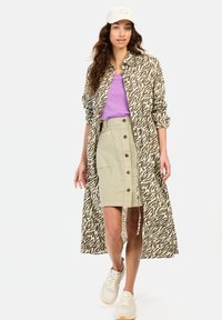 Lange mouwen, dierenprint shirt in olijfgroen en beige, gecombineerd met een lavendeltop en een beige rok met knopen, afgemaakt met witte sneakers.