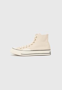 Beige high-top sneakers made of canvas, featuring a rubber toe cap, white sole, black stripe, and gold eyelets for laces.