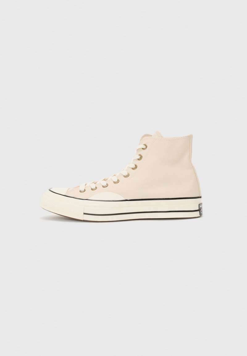 Beige high-top sneakers made of canvas, featuring a rubber toe cap, white sole, black stripe, and gold eyelets for laces.