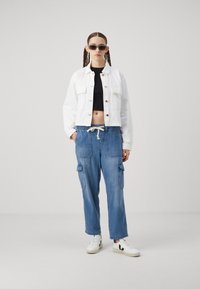 Vit kortärmad denimjacka, svart crop top, blå cargobyxor med fickor och vita sneakers. Modellen bär bruna solglasögon.