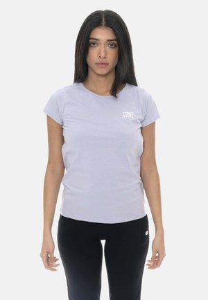 Donna con capelli lunghi e scuri che indossa una maglietta viola chiaro a maniche corte e pantaloni neri, in piedi davanti a uno sfondo bianco semplice.