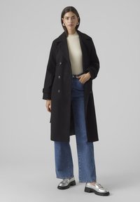 Cappotto doppiopetto nero con ampi revers, indossato sopra un maglione a coste crema e jeans flare blu a vita alta, accentuato da scarpe argentate.