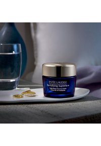Blå glaskrukke med Estée Lauder Revitalizing Supreme+ creme med et gyldent låg, ved siden af en hvid tallerken der bærer tre gyldne ringe og et vandglas.