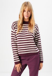 Young woman with long blonde hair wearing a burgundy and white striped knit sweater and matching burgundy pants against a white background.