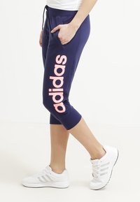 Tummansinisiä lyhyitä jogger-housuja, joissa on vaaleanpunainen "adidas"-logo vasemmassa lahkeessa, sivutaskut ja narukaulus. Yhdistetty valkoisiin tennareihin.