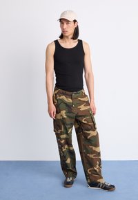 Pantaloni cargo mimetici verdi e marroni con tasche, abbinati a una canotta nera e un cappello beige. Scarpe da ginnastica nere con lacci bianchi, in posizione eretta.