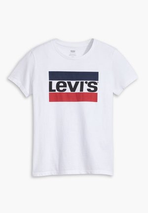 Person iført en simpel hvid t-shirt med korte ærmer og lyseblå denimjeans, vist bagfra mod en ensfarvet baggrund.