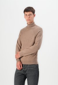 Pull col roulé beige avec accents côtelés sur les épaules et les poignets, fabriqué dans un matériau doux, associé à un jean en denim gris.