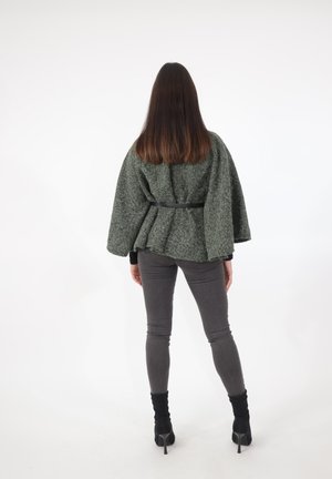 Frau steht mit dem Gesicht abgewandt, trägt einen grauen, strukturierten Umhang mit Gürtel, eng anliegende graue Hose und schwarze, knöchelhohe Stiefeletten mit hohen Absätzen vor weißem Hintergrund.