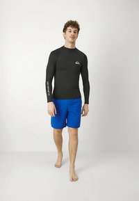 Camisola de proteção contra o sol preta de manga comprida com o logótipo Quiksilver, combinada com calções de banho azuis. Textura suave, corte atlético e design leve.