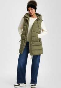 Padded oliwkowa kamizelka z kapturem, z zamkiem błyskawicznym, bocznymi kieszeniami i wiązanym dołem, noszona na białym, textured swetrze i szerokich dżinsach.