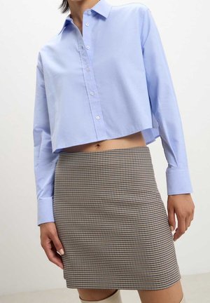 Personne portant une chemise courte bleu clair à boutons et une jupe taille haute marron et blanche à motif pied-de-poule sur un fond uni.