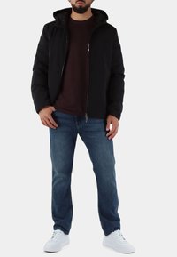 Zwarte jas met rits en capuchon, bordeauxrode shirt eronder, blauwe spijkerbroek en witte sneakers; casual laagjes met strakke texturen.