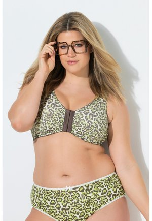 Kobieta o kobiecych kształtach nosząca zielony i czarny biustonosz w panterkę i dopasowane majtki, poprawiająca duże okulary w oprawkach typu 'tortoiseshell', na białym tle.