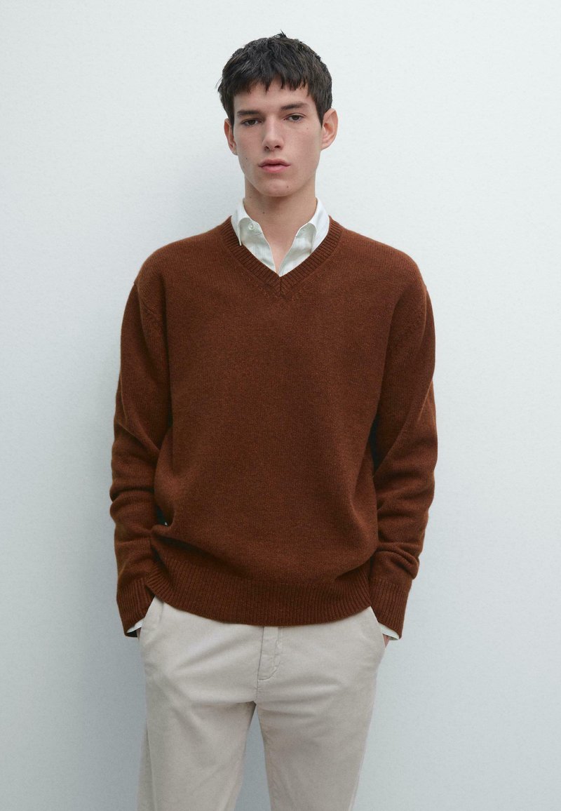 Jonge man draagt een bruine trui met V-hals over een wit shirt en beige broek, staand tegen een effen lichtgrijze achtergrond.