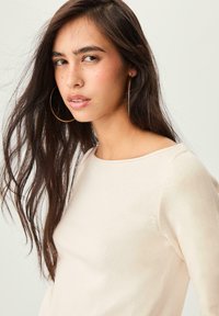 Cremefarbener Pullover mit langen Ärmeln und rundem Halsausschnitt, aus weichem Stoff gefertigt. Das Model hat lange, dunkle Haare und trägt große Creolen.