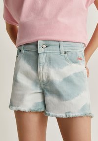 Shorts de denim azul claro con patrón de tie-dye blanco, dobladillo deshilachado y logo bordado rojo en el bolsillo delantero. Cierre con botón plateado.