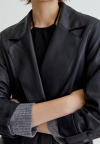 Blazer en cuir noir avec revers crantés, doté d'un poignet en maille grise douce. Texture lisse et coupe ajustée, avec des accents de matériel minimalistes.