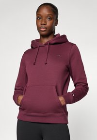 Hoodie bordeaux avec une poche frontale, capuche à cordon de serrage et poignets côtelés. Présente un petit logo sur la poitrine et une texture douce.